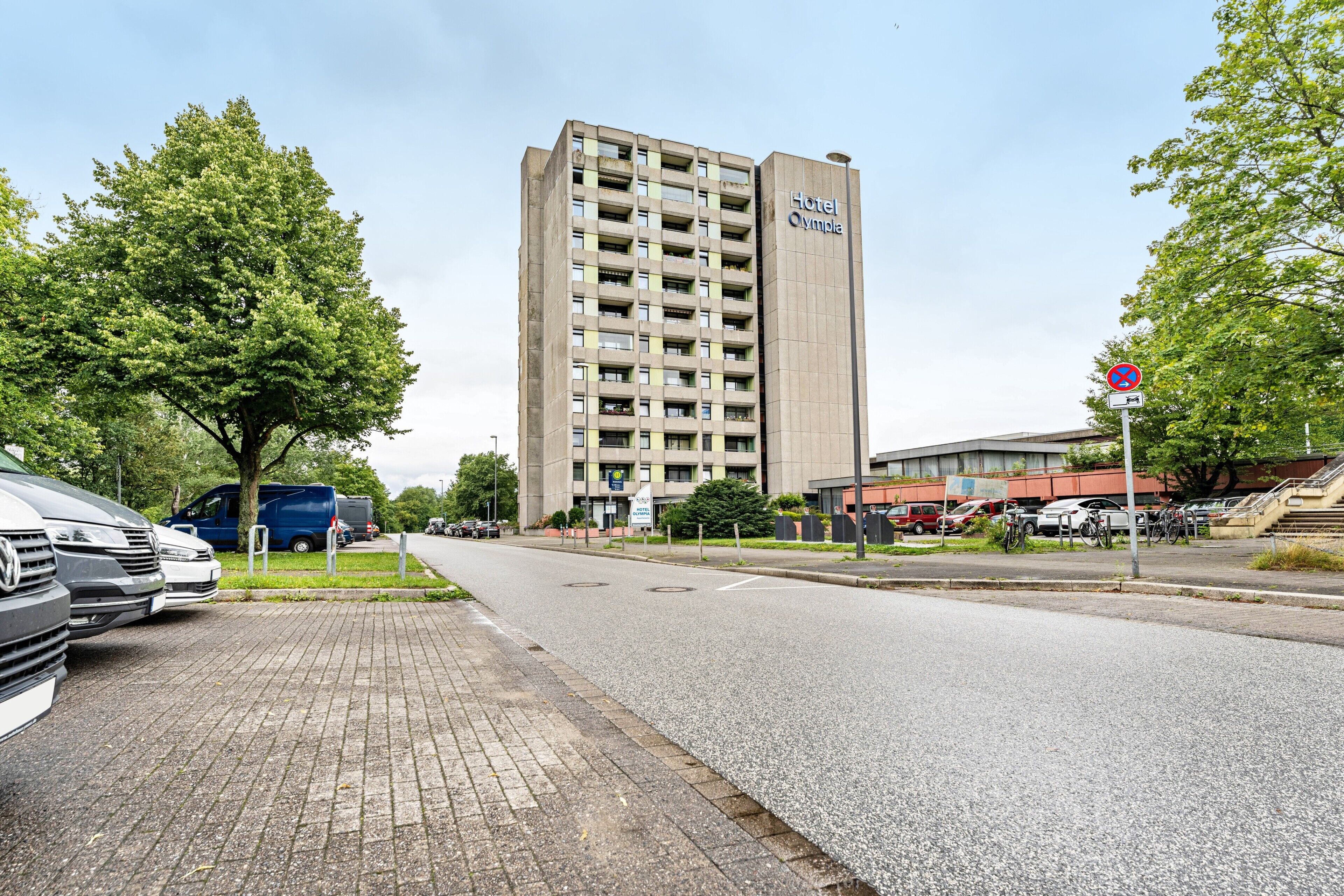 Ein hohes Hotelgebäude mit dem Schriftzug 'Hotel Olympia', einem gepflasterten Bürgersteig, geparkten Autos und einem Verbotsschild.