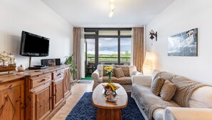 A living room with a TV, wooden cabinet, sofa, coffee table, and a view through large windows.