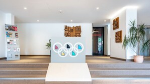 A reception area with a modern design, featuring a central podium with Olympic rings, a wall-mounted wooden art piece, and a potted plant.