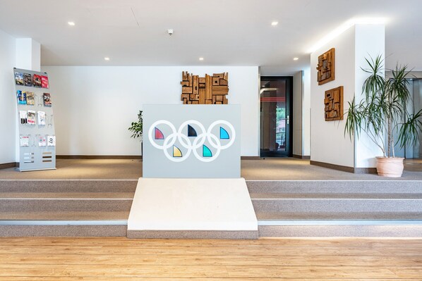 A reception area with a modern design, featuring a central podium with Olympic rings, a wall-mounted wooden art piece, and a potted plant.