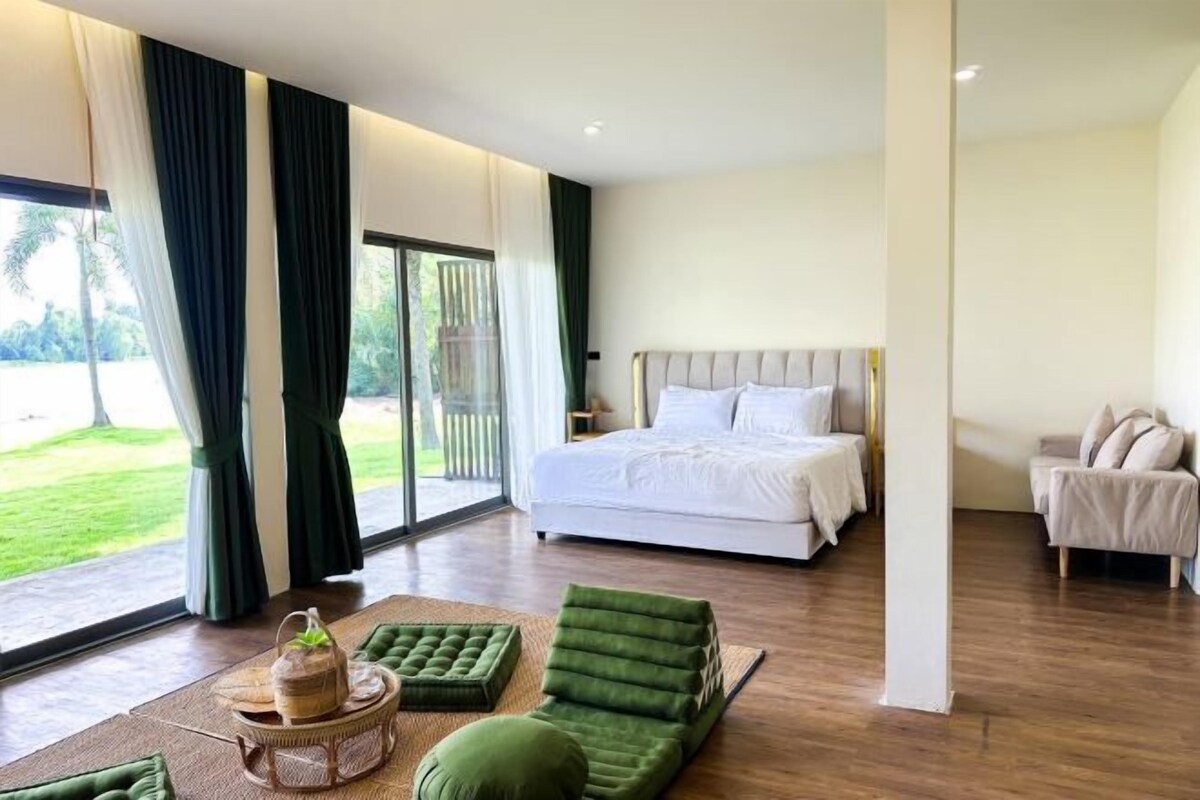 A spacious bedroom with a large bed, a seating area with green cushions, and a view of a lush garden through the sliding glass door.