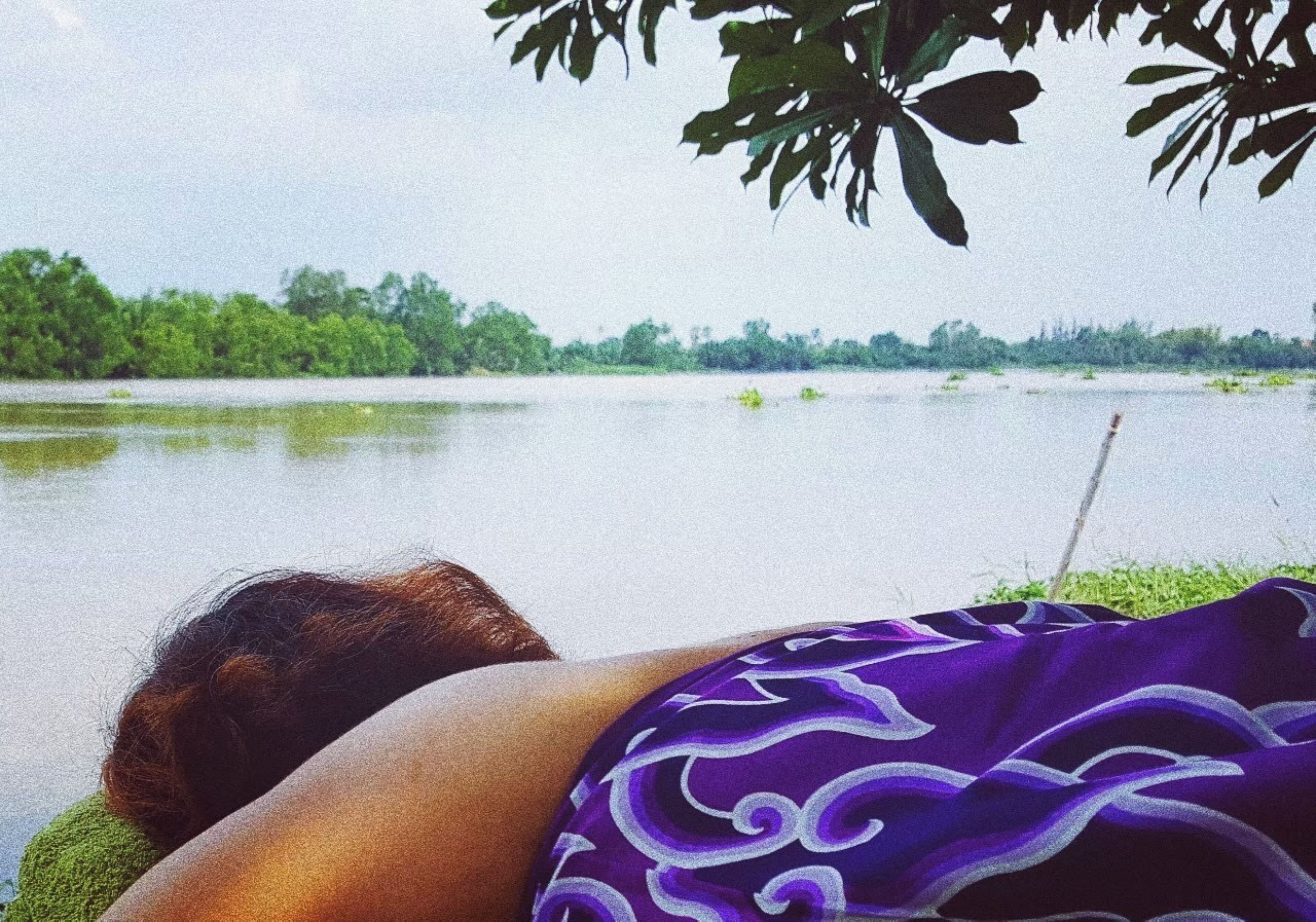 A person lying on a bed with a view of a lake and trees in the background.