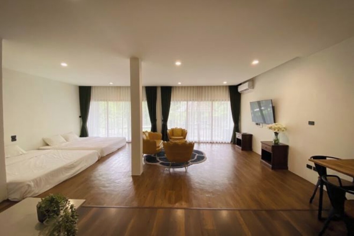 A modern hotel room with a wooden floor, a bed, two yellow armchairs, a small table, a TV, and a window with curtains.