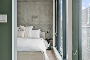 Une chambre avec une grande fenêtre, un lit recouvert de draps blancs, une table de chevet avec une lampe et un mur en béton.