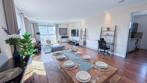 Uma sala de estar espaçosa com uma grande mesa de jantar posta para seis pessoas, um sofá, uma TV e uma área de cozinha.