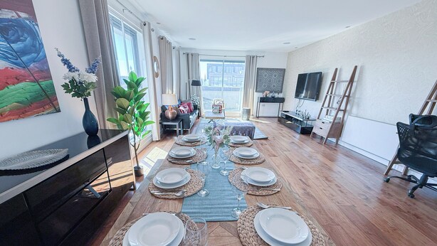 Uma sala de jantar com uma mesa comprida posta para seis pessoas, um vaso com planta e um quadro na parede.