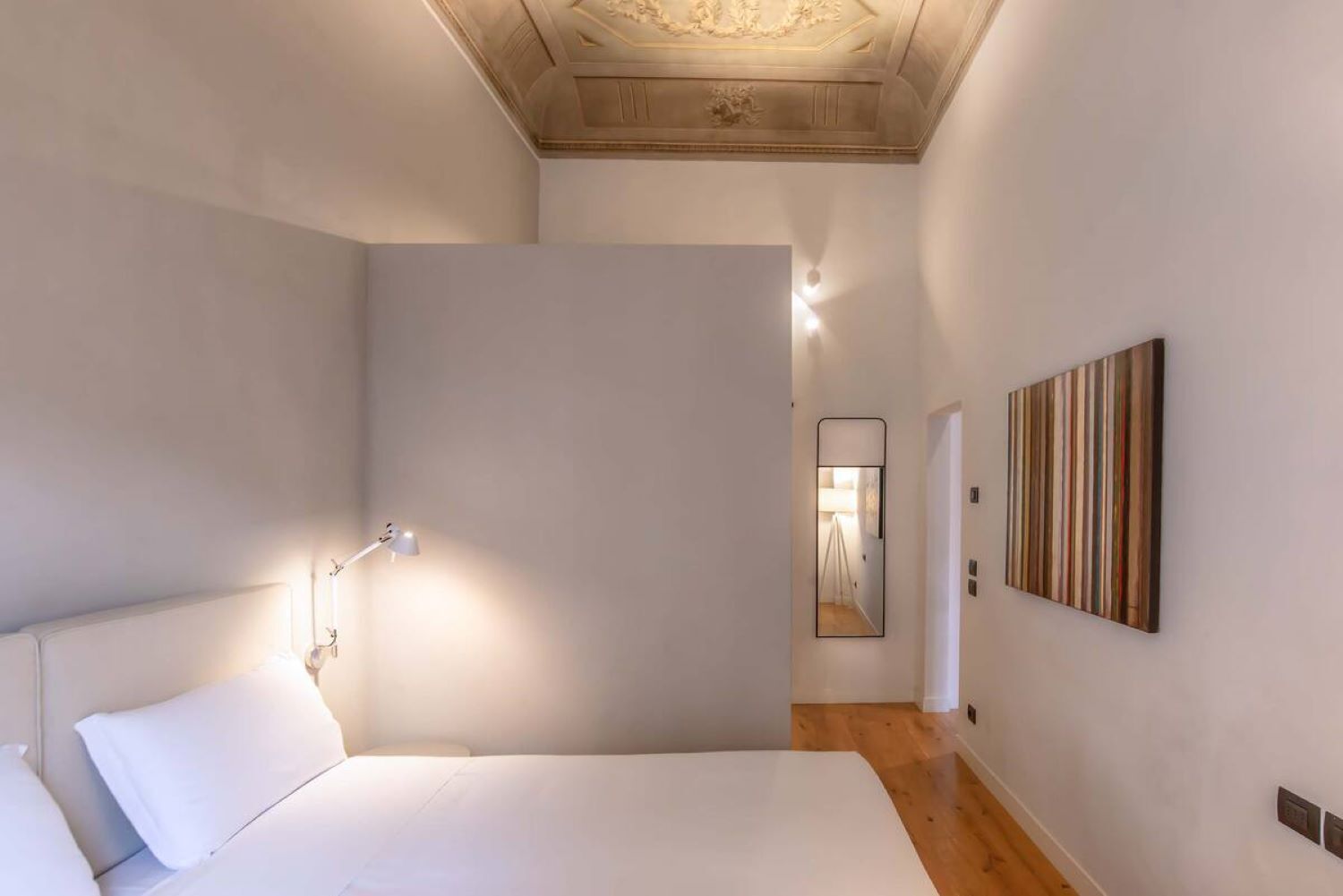 Une chambre à coucher avec un lit, une lampe de chevet, un plancher en bois, un miroir et une œuvre d’art en bois accrochée au mur.