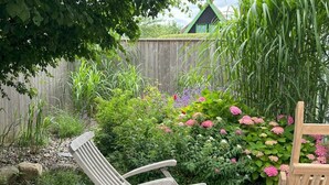Un giardino con un patio in legno, due sedie di legno e diverse piante fiorite.