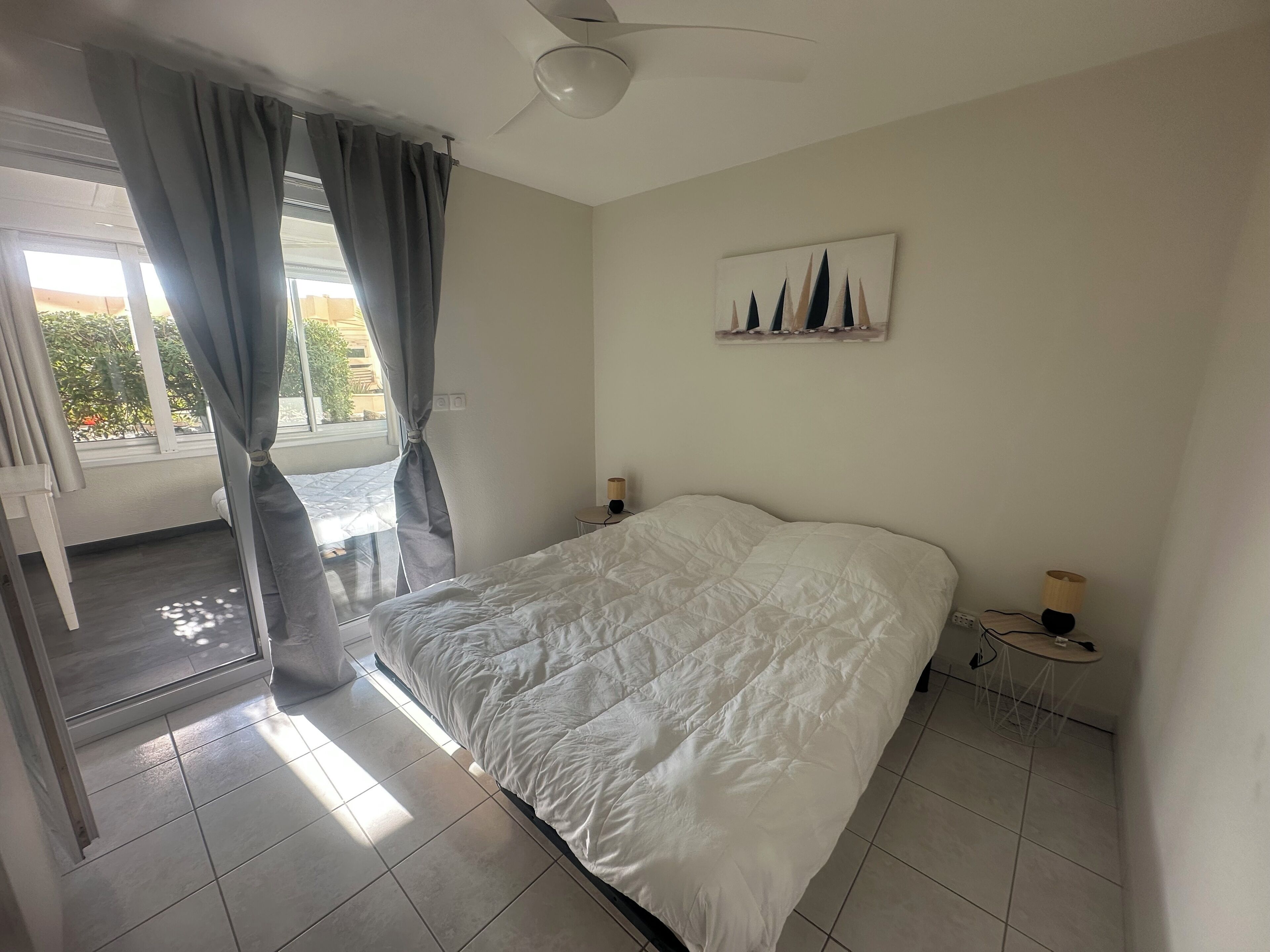 Une chambre à coucher comprenant un lit, des rideaux, un ventilateur de plafond, une table de chevet avec une lampe et un tableau accroché au mur.