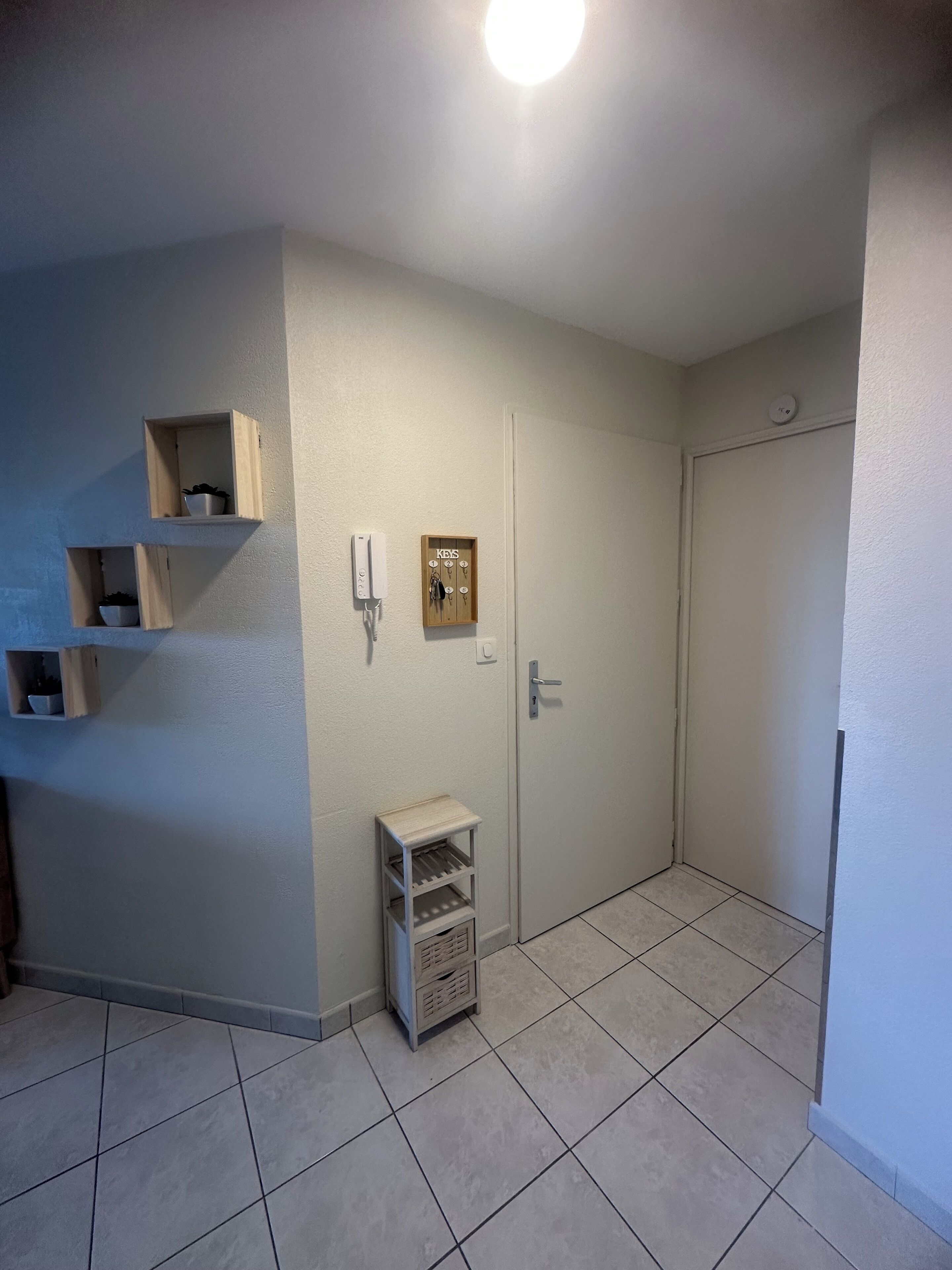 A small, clean room with a tiled floor, a door, a wall-mounted phone, and a key rack.
