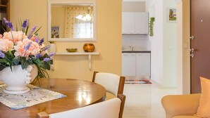 Un coin repas avec une table ronde, des chaises, un vase de fleurs, un miroir et une vue sur la cuisine.