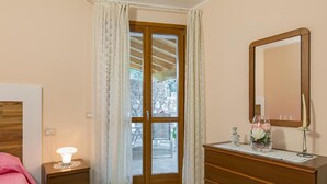 Une chambre avec une commode en bois, une table de chevet avec une lampe, un miroir et une vue sur l’extérieur, accessible par une porte vitrée.