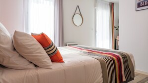 Un dormitorio con cama, almohadas, una manta a rayas, un espejo redondo y una ventana con cortinas.