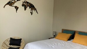Quarto com uma cama grande, duas cadeiras de vime, uma mesa redonda pequena, luminária pendente e obras de arte na parede.