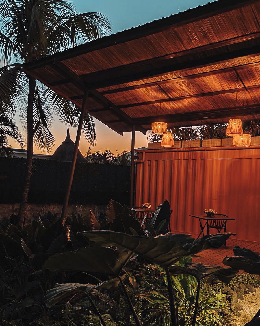 Ein Terrassenbereich im Freien mit überdachter Sitzecke, einem Tisch und Stühlen, sowie Palmen und einem Gebäude im Hintergrund.