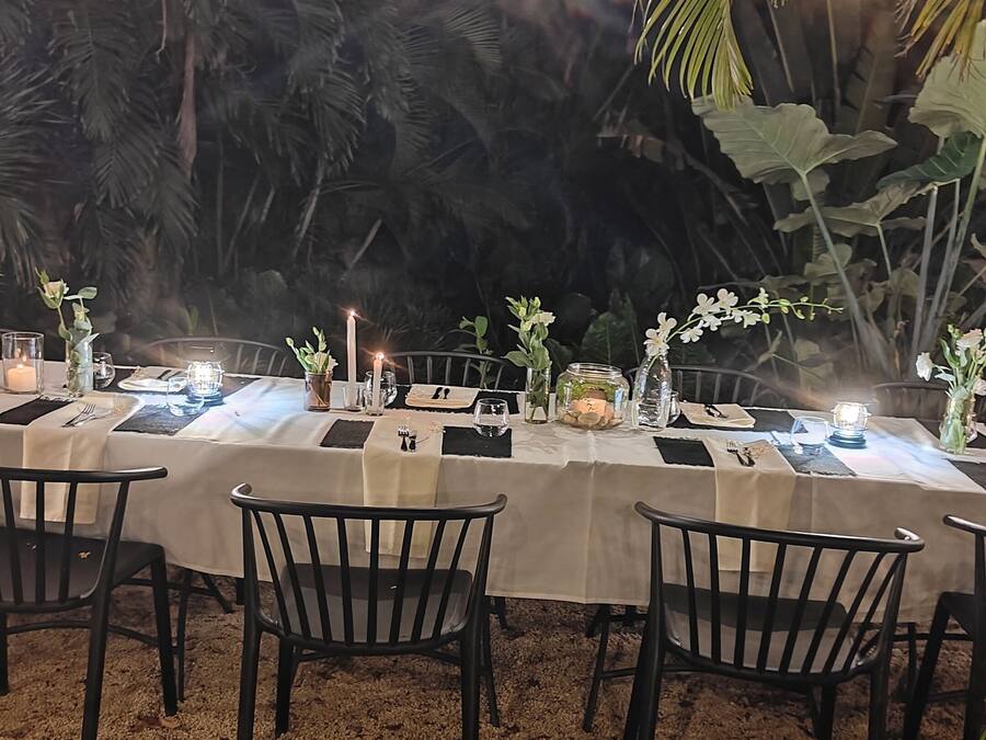 An outdoor dining area with tables set for a meal, surrounded by palm trees and a gravel floor.