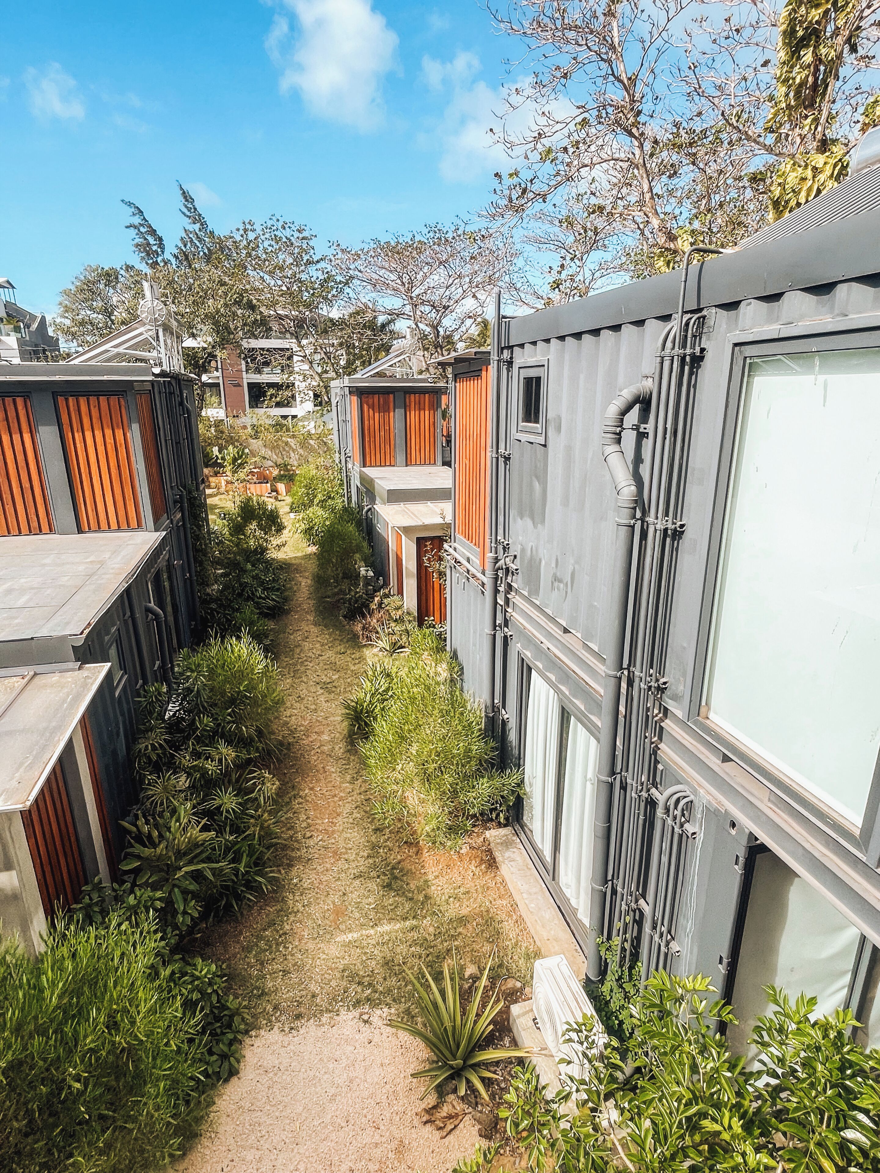 Un sendero estrecho entre edificios modulares con contraventanas de madera, rodeado de follaje.