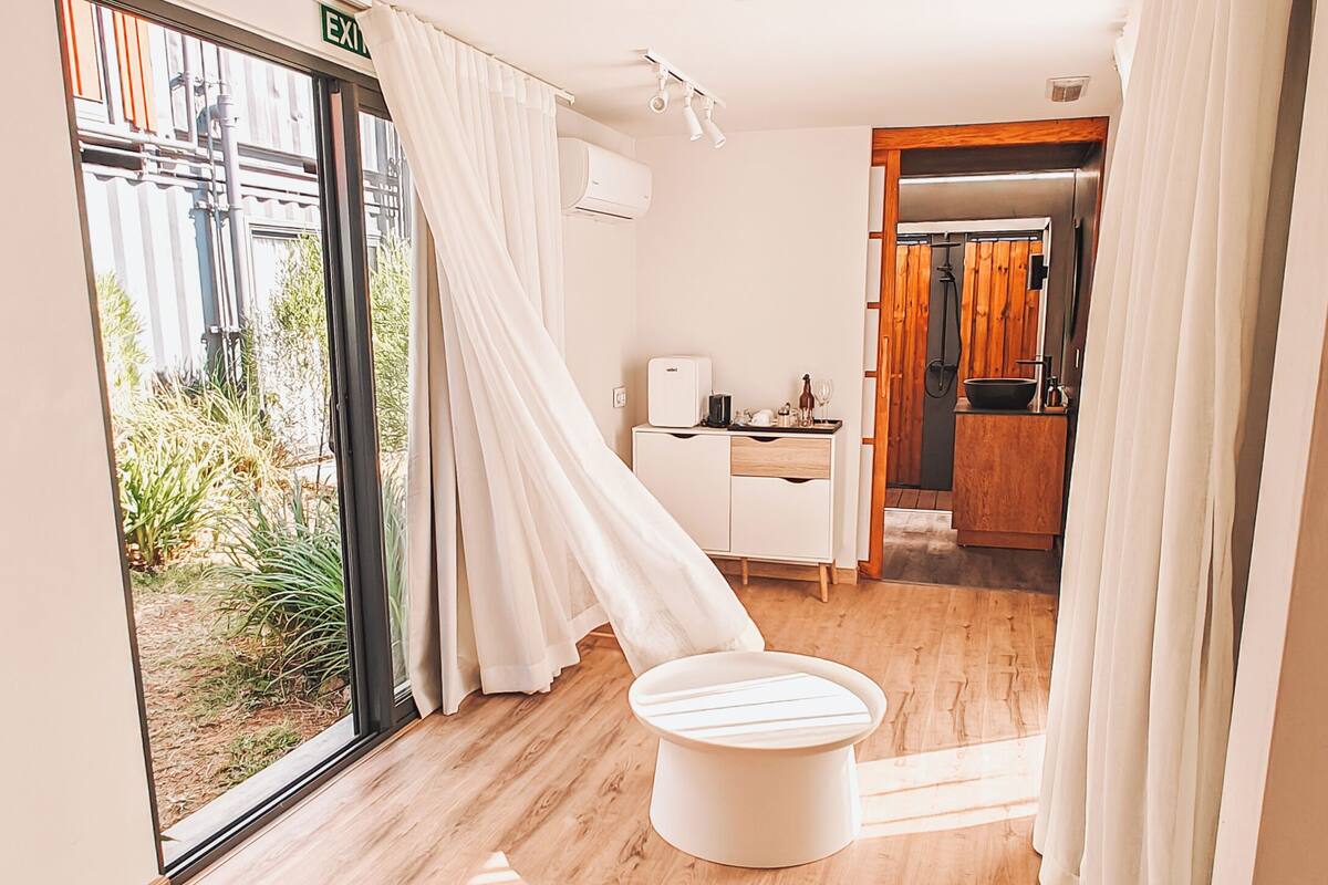 A modern hotel room with a wooden floor, a round white table, a small white cabinet, and a view of greenery outside.