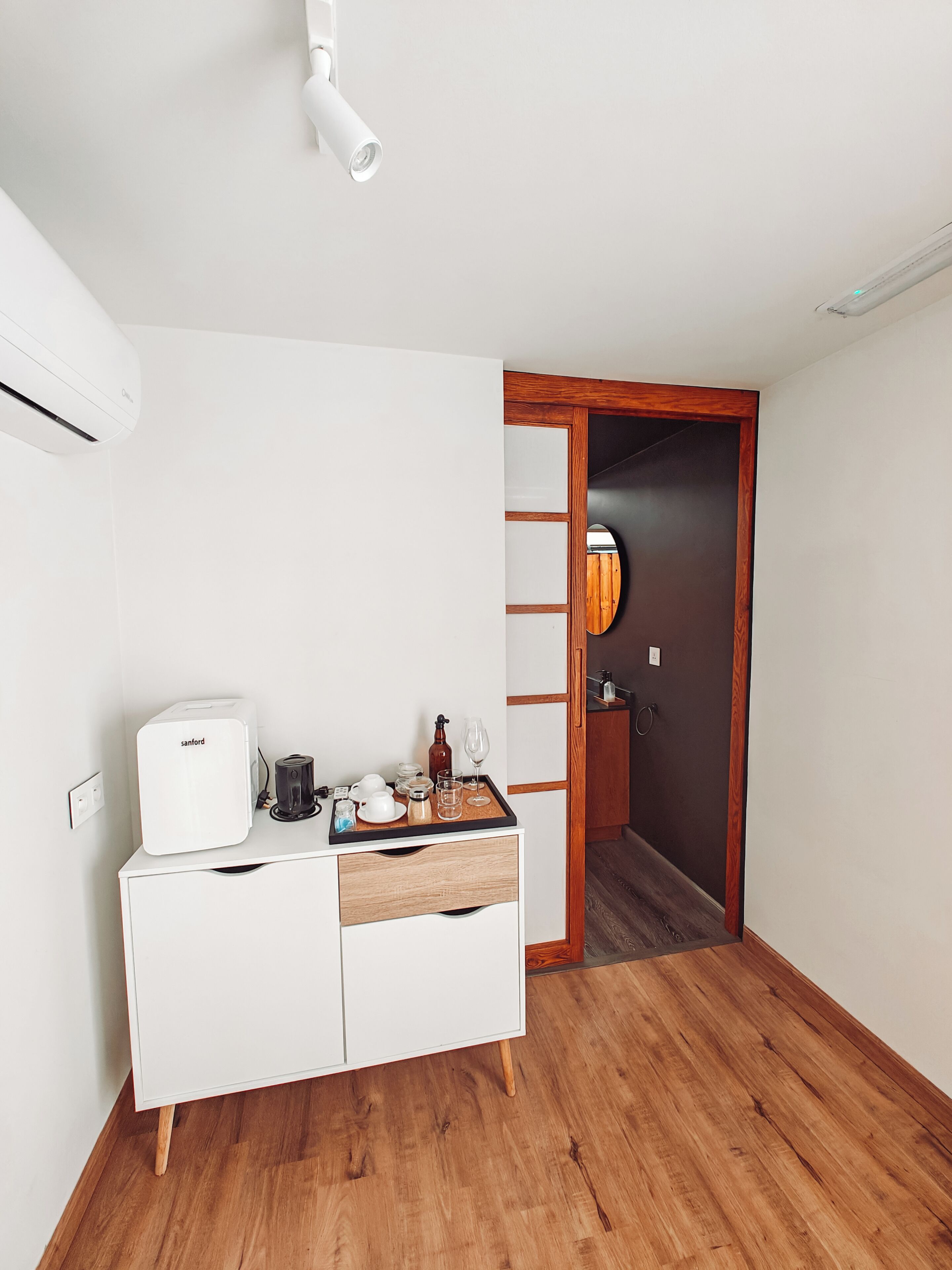 Una habitación pequeña y limpia con un armario blanco, suelo de madera, una lámpara en el techo y una puerta que da a otra habitación.