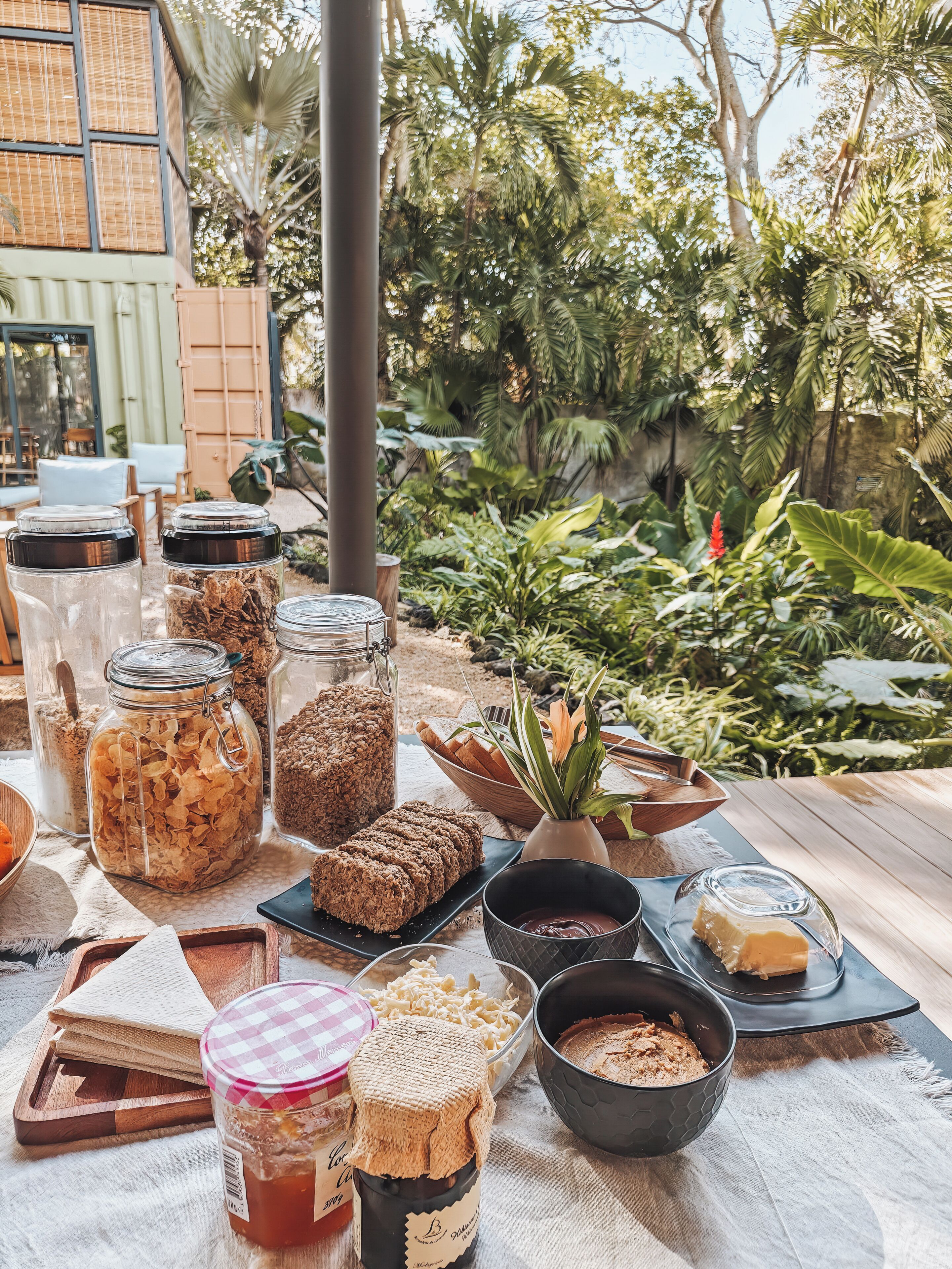 An outdoor dining area with various food items, including jars of granola and bread, set against a backdrop of lush greenery and a building.