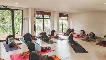 Una clase de yoga en una habitación con ventanas amplias, una puerta con la inscripción 'EXIT' y suelo de madera.