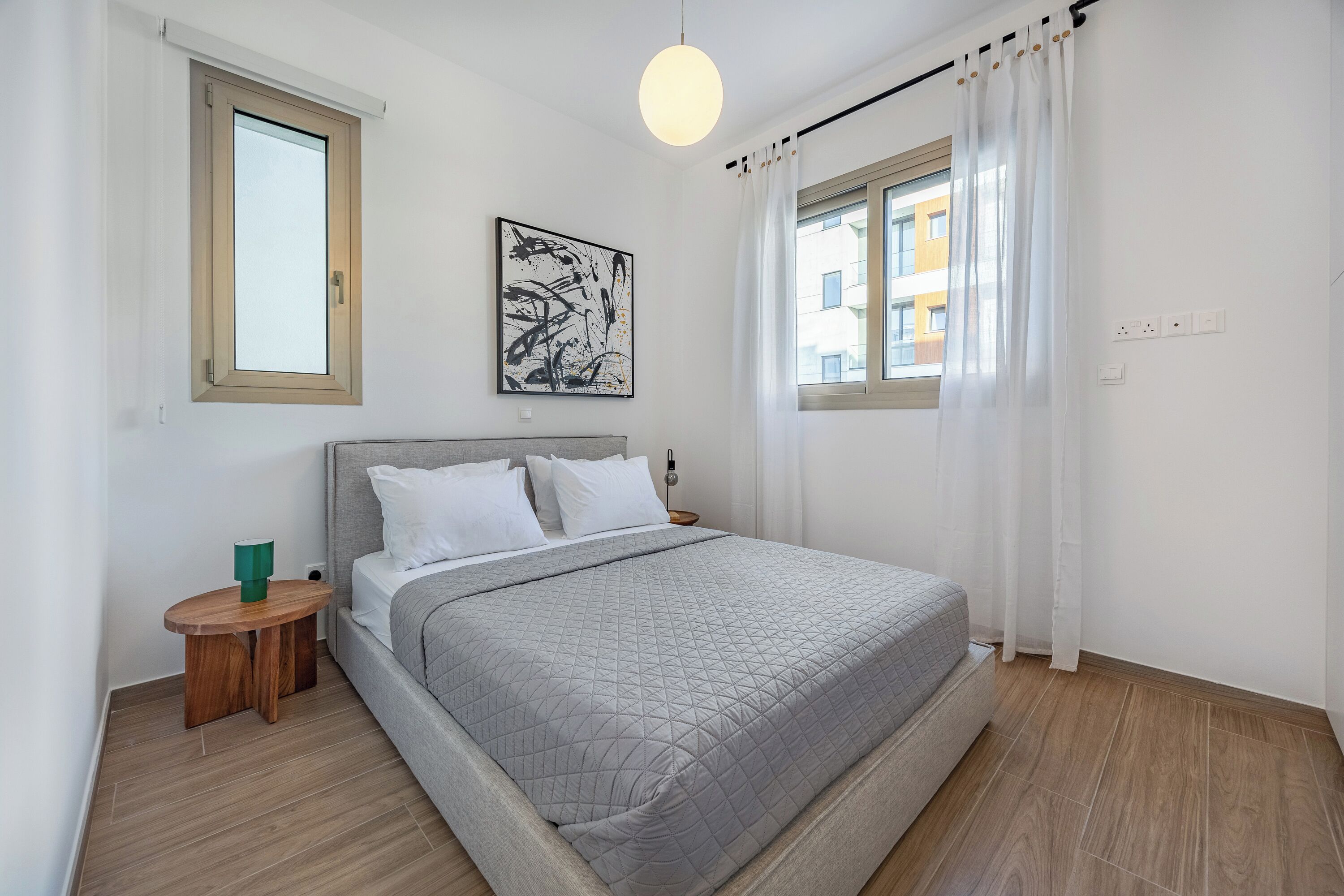 Un dormitorio moderno con una cama grande, una mesita de noche, una lámpara de pared y un ventanal con cortinas.