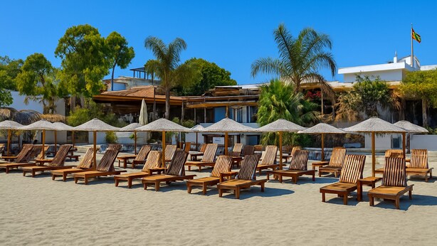 A beachside hotel with rows of wooden lounge chairs and umbrellas, palm trees, and a clear blue sky. - Mulberry Court Premium Apartments (Limassol)
