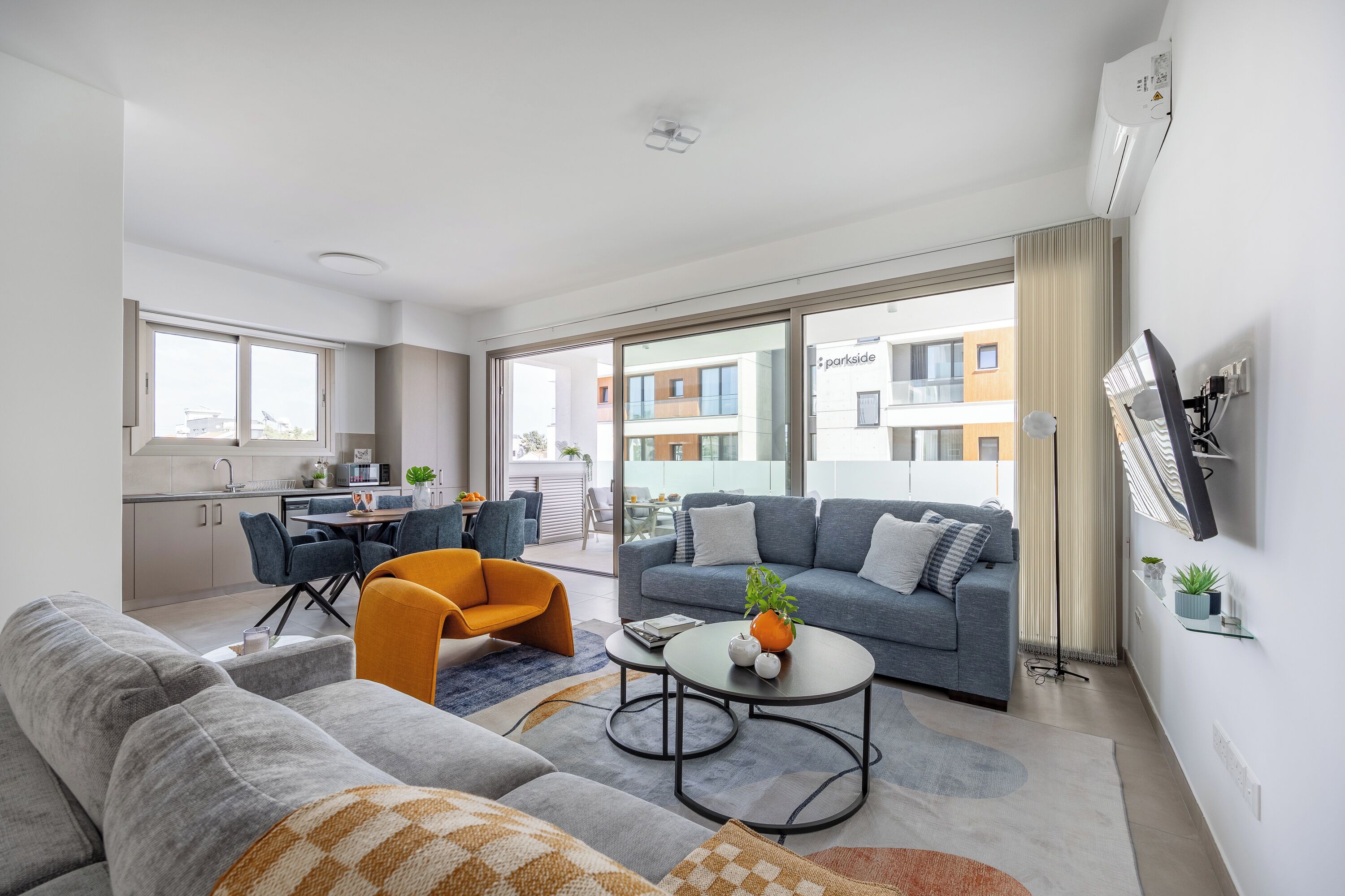 Een moderne woonkamer met een grijze bank, een oranje fauteuil en een grijze divan. Er is een eethoek met een tafel en stoelen, en een keuken op de achtergrond.