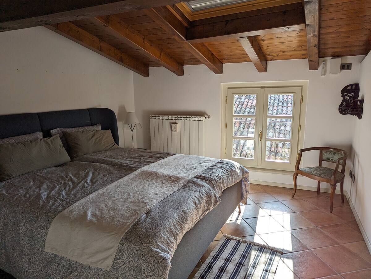 A bedroom with a large bed, wooden beams on the ceiling, a radiator, a window with a view, and a chair.
