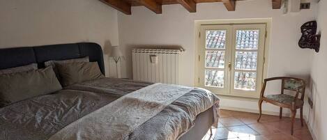 A bedroom with a large bed, wooden beams on the ceiling, a radiator, a window with a view, and a chair.