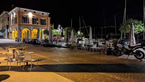 Een terras met tafels en stoelen, een motorfiets geparkeerd, en een gebouw met vlaggen op de achtergrond.