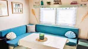 A cozy room with a blue sofa, a small table with a patterned tablecloth, and a window with blinds. - Air-conditioned mobile home, 5-minute walk from Lac de Biscarrosse "Menthe à l'eau" lake (Biscarrosse)