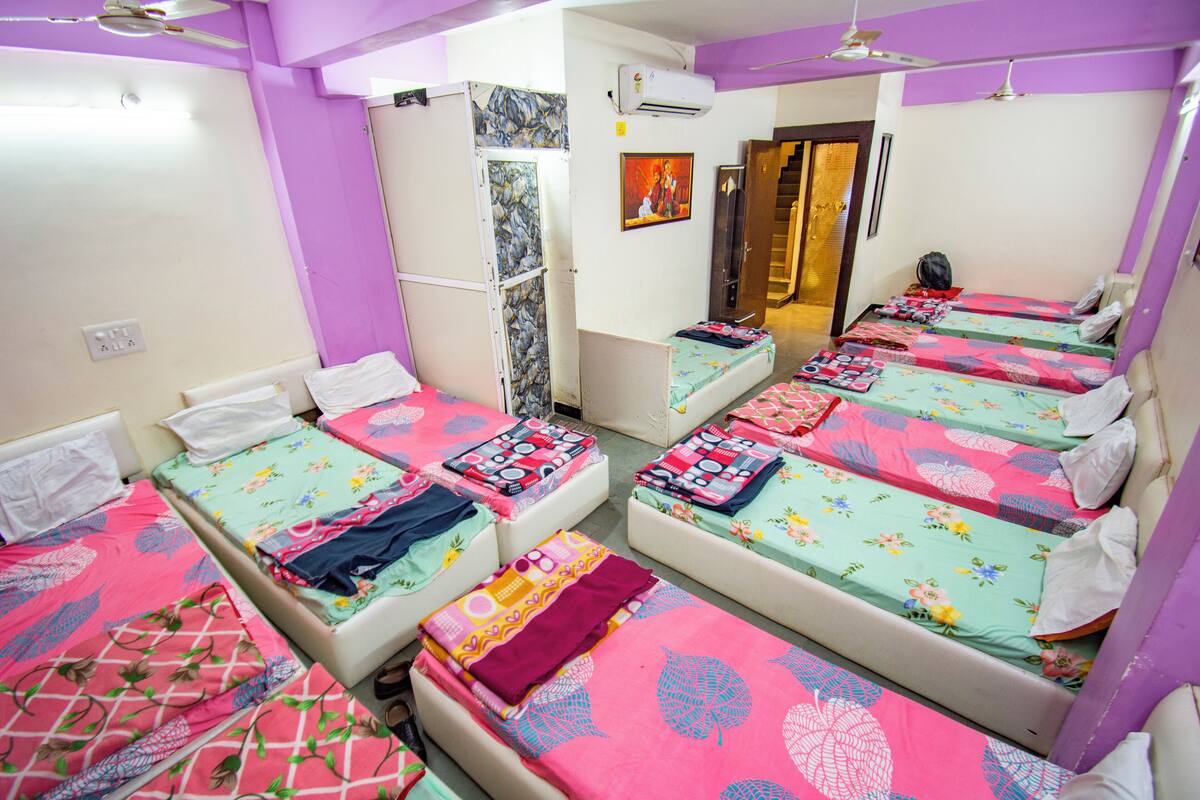 A room with multiple beds, each with colorful bedding. There are ceiling fans, an air conditioning unit, and a door leading to another room.