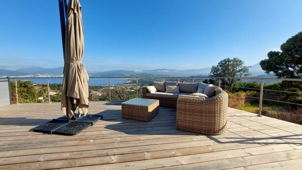Una terraza de madera con mobiliario de mimbre y una sombrilla, con vistas a un paisaje pintoresco con montañas y una masa de agua.