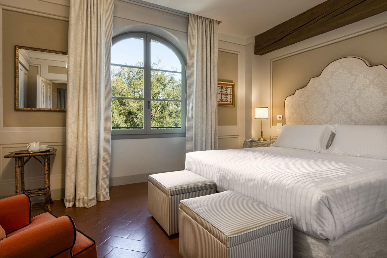 Een hotelkamer met een groot bed, twee poufs, een klein tafeltje met een theepot, een stoel en een raam met gordijnen.