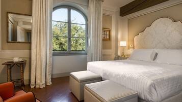 Een hotelkamer met een groot bed, twee poufs, een klein tafeltje met een theepot, een stoel en een raam met gordijnen.