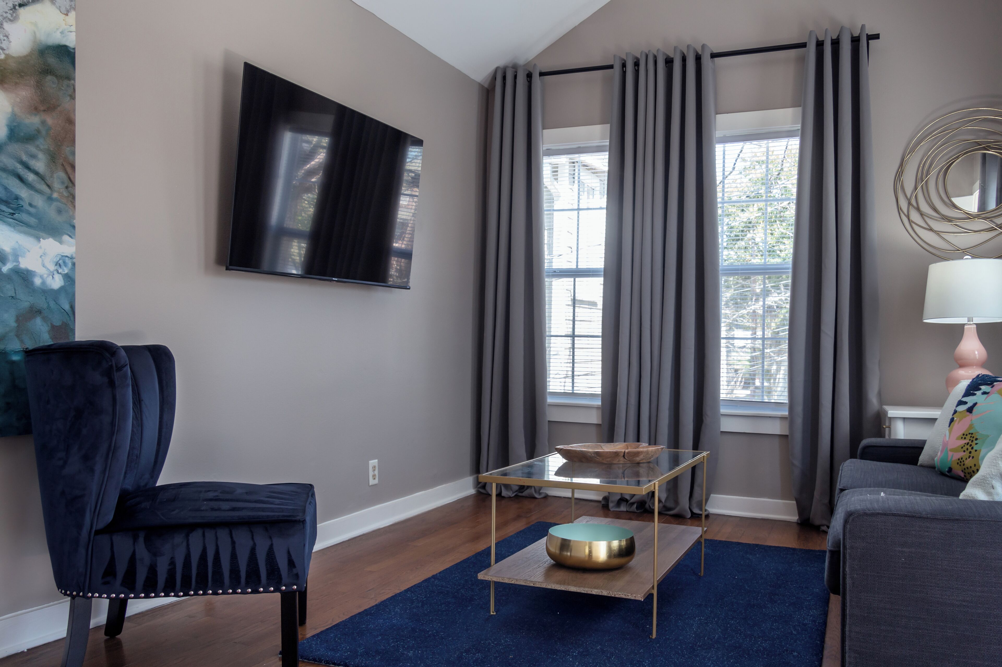 Een moderne woonkamer met een donkerblauwe fauteuil, een glazen salontafel met een gouden schaal, een grijze bank en een aan de muur bevestigde televisie.