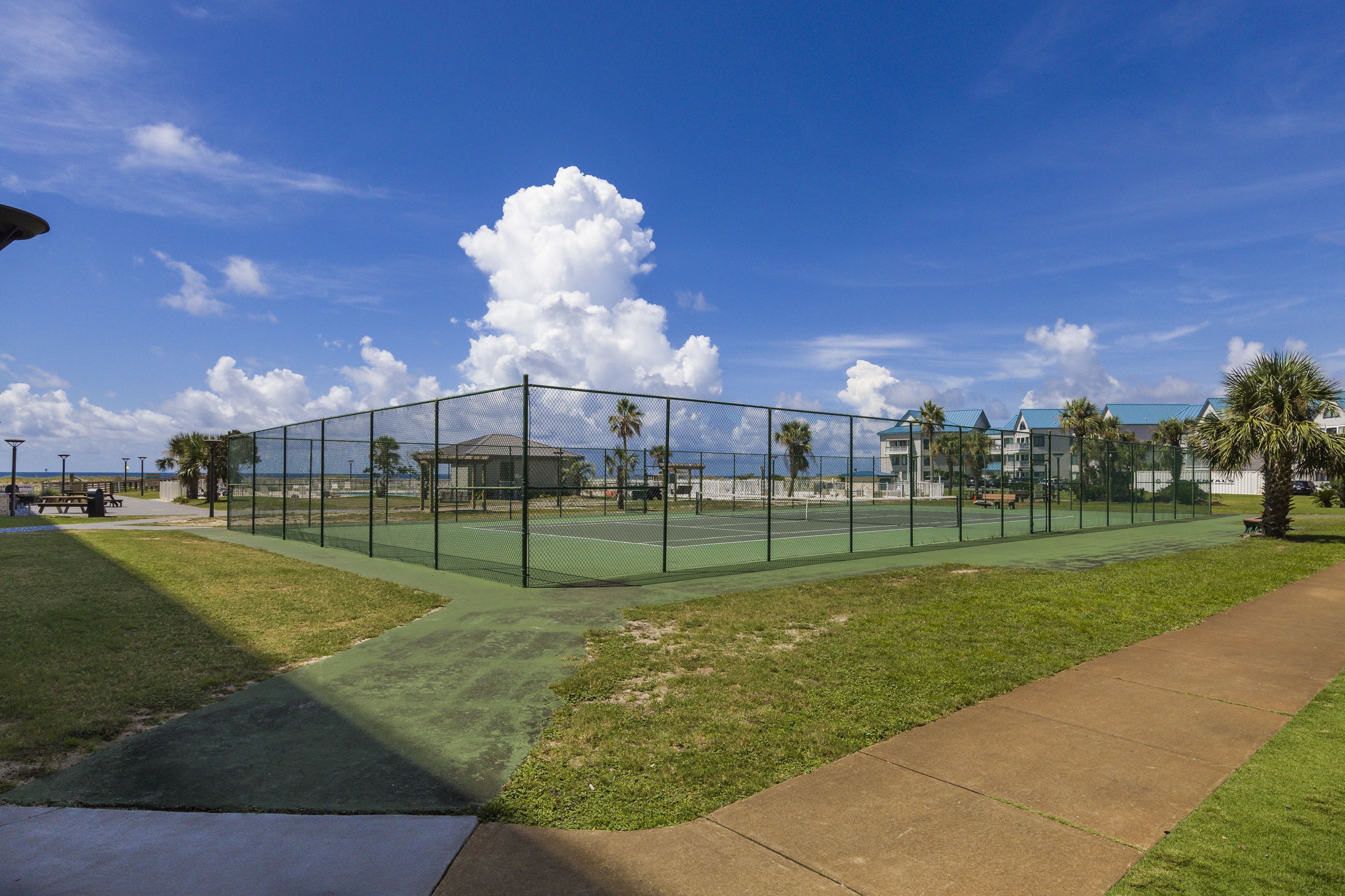 A tennis court with a fence, a grassy area, and a sidewalk.