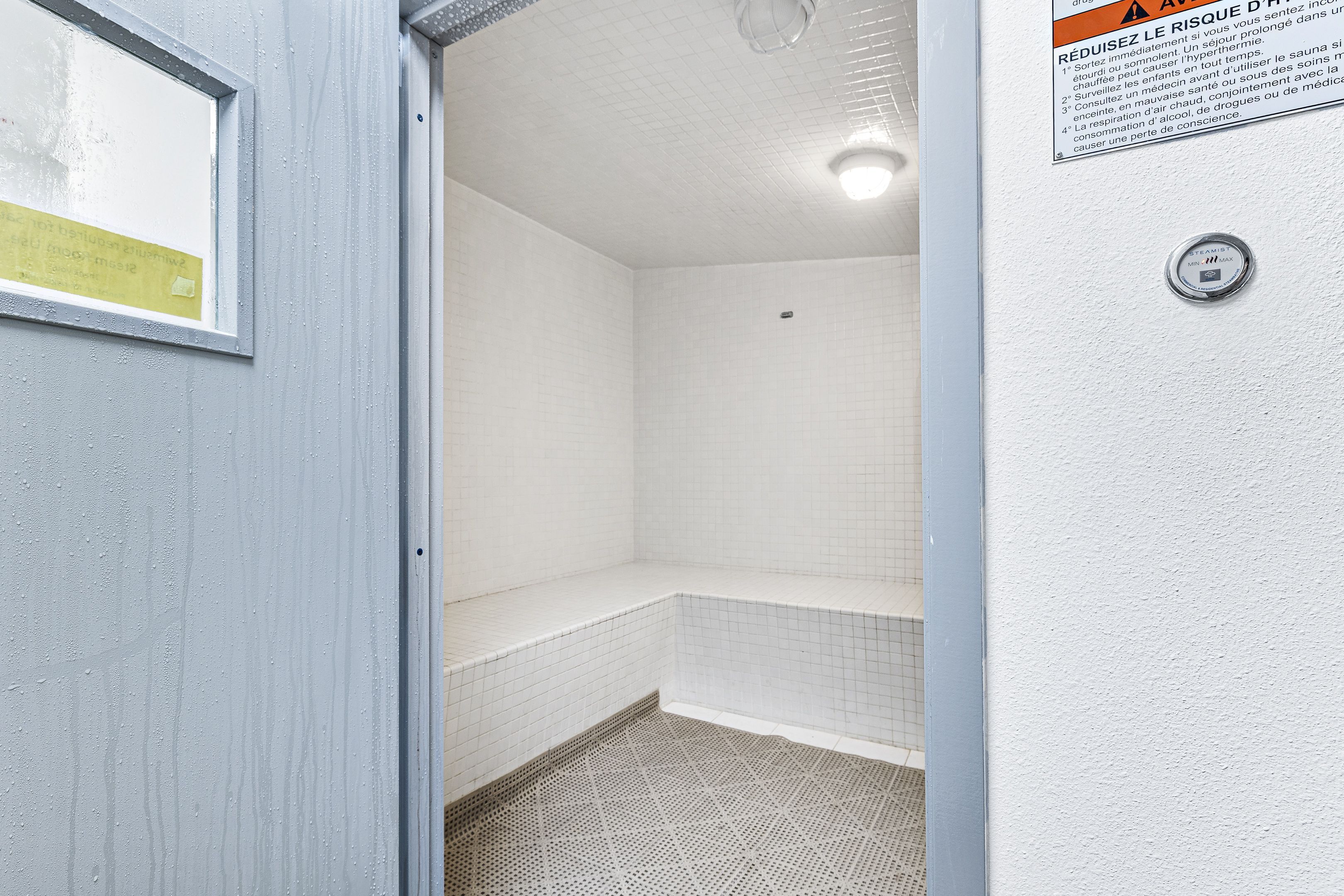 Une salle de bain avec des murs et un sol recouverts de carreaux blancs, un espace douche et une porte dotée d’une fenêtre.