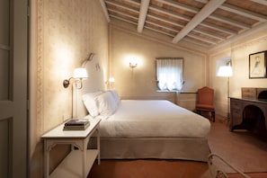 A hotel room with a bed, bedside table, lamp, chair, desk, and a fireplace. - My Exclusive Italy - Classic Suite in a Luxury Wine Estate (Rignano sull'Arno)