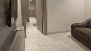 Un pasillo con piso de mármol que conduce a un baño con lavamanos y espejo. Se ve un sofá marrón a la derecha.