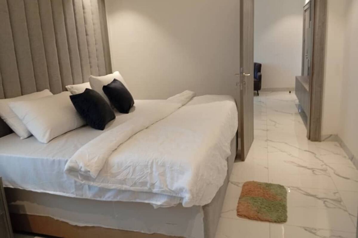 Um quarto com cama, almofadas, mesa de cabeceira e uma porta que dá para outro cômodo.