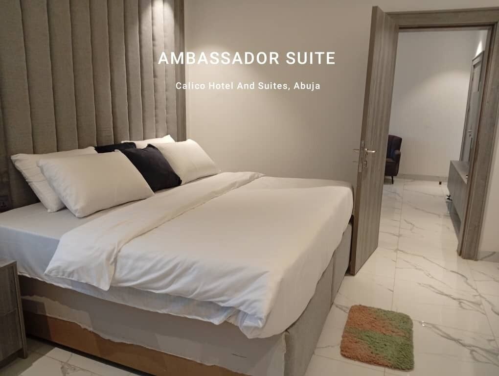 An Ambassador Suite at Calico Hotel And Suites, Abuja. The suite features a large bed with white bedding, a wooden headboard, and a marble floor.