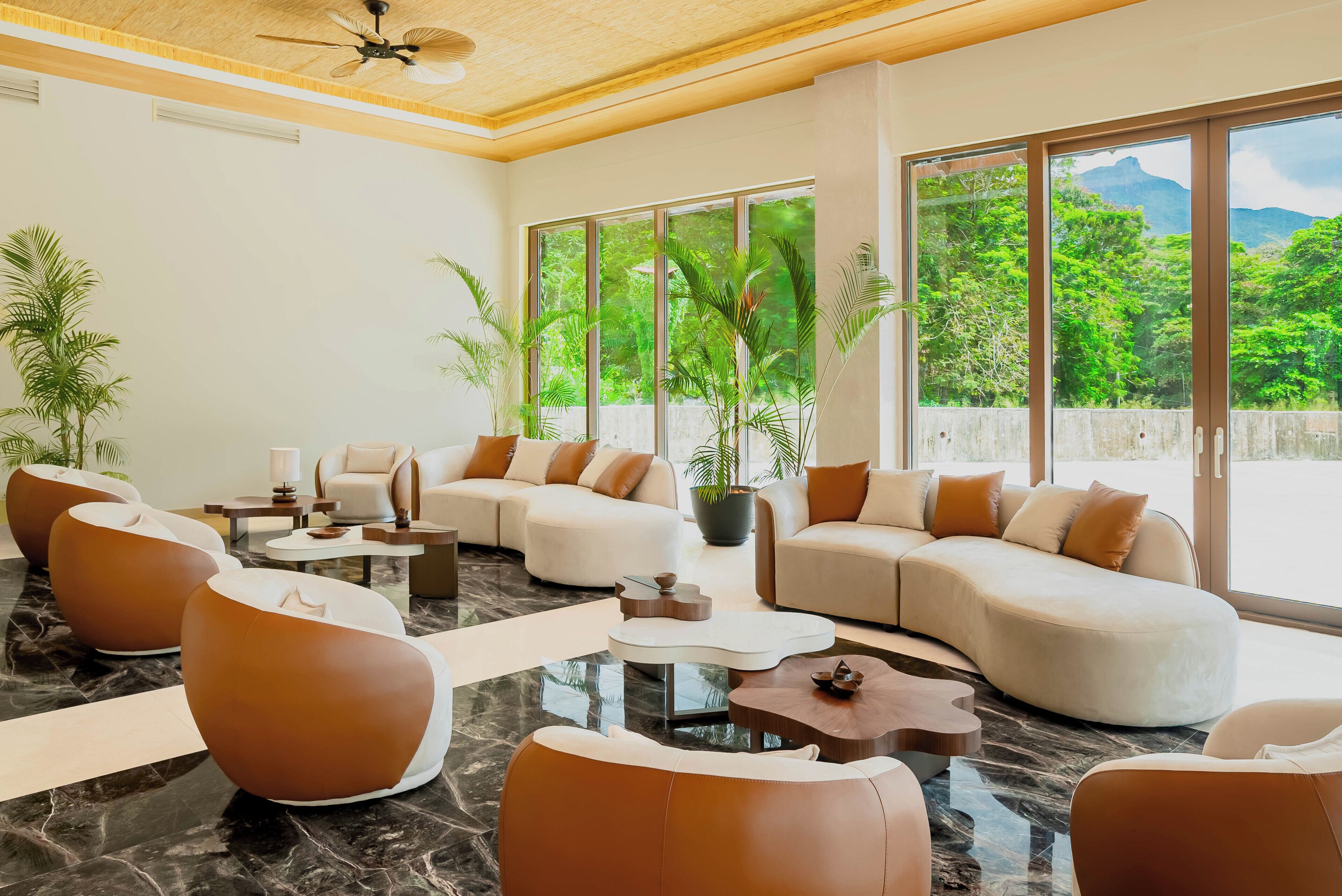 A spacious living area with modern furniture, large windows offering a view of lush greenery and mountains, and a ceiling fan.