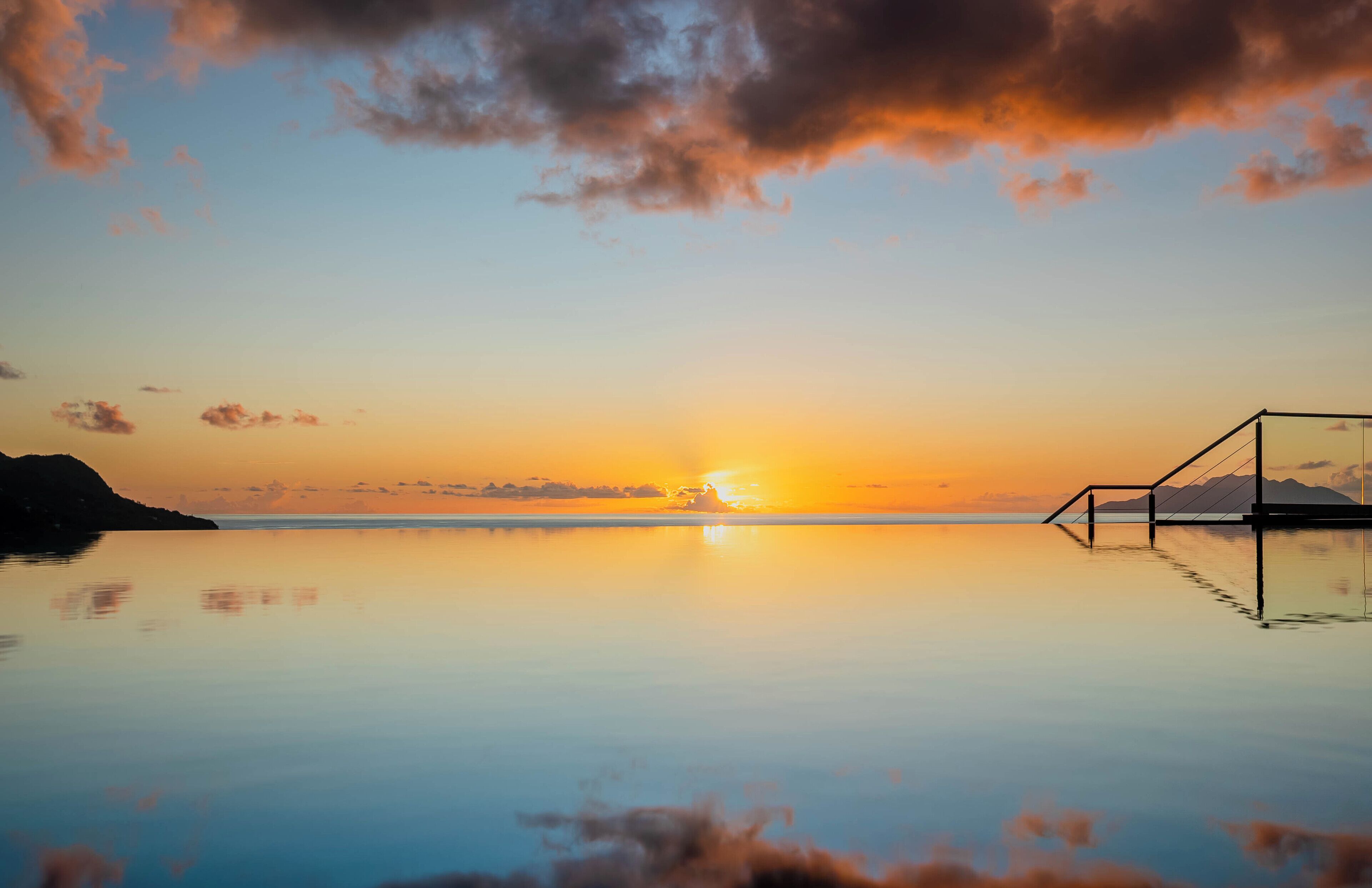 A tranquil sunset over a calm body of water with a glass-bottomed infinity pool and a simple metal railing.