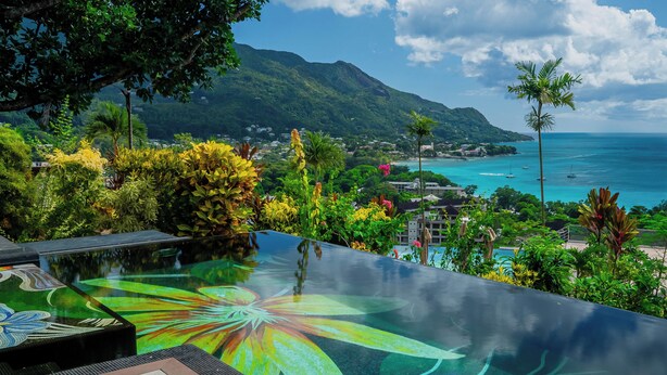 Une piscine décorée de motifs floraux tropicaux, entourée d’une végétation luxuriante et offrant une vue sur une ville côtière, avec des montagnes en arrière-plan.