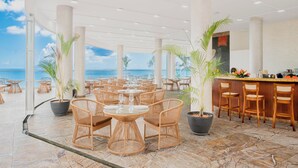 Un restaurant situé en bord de mer, avec des tables rondes et des chaises en osier, de grandes plantes en pot et une vue sur l’océan.