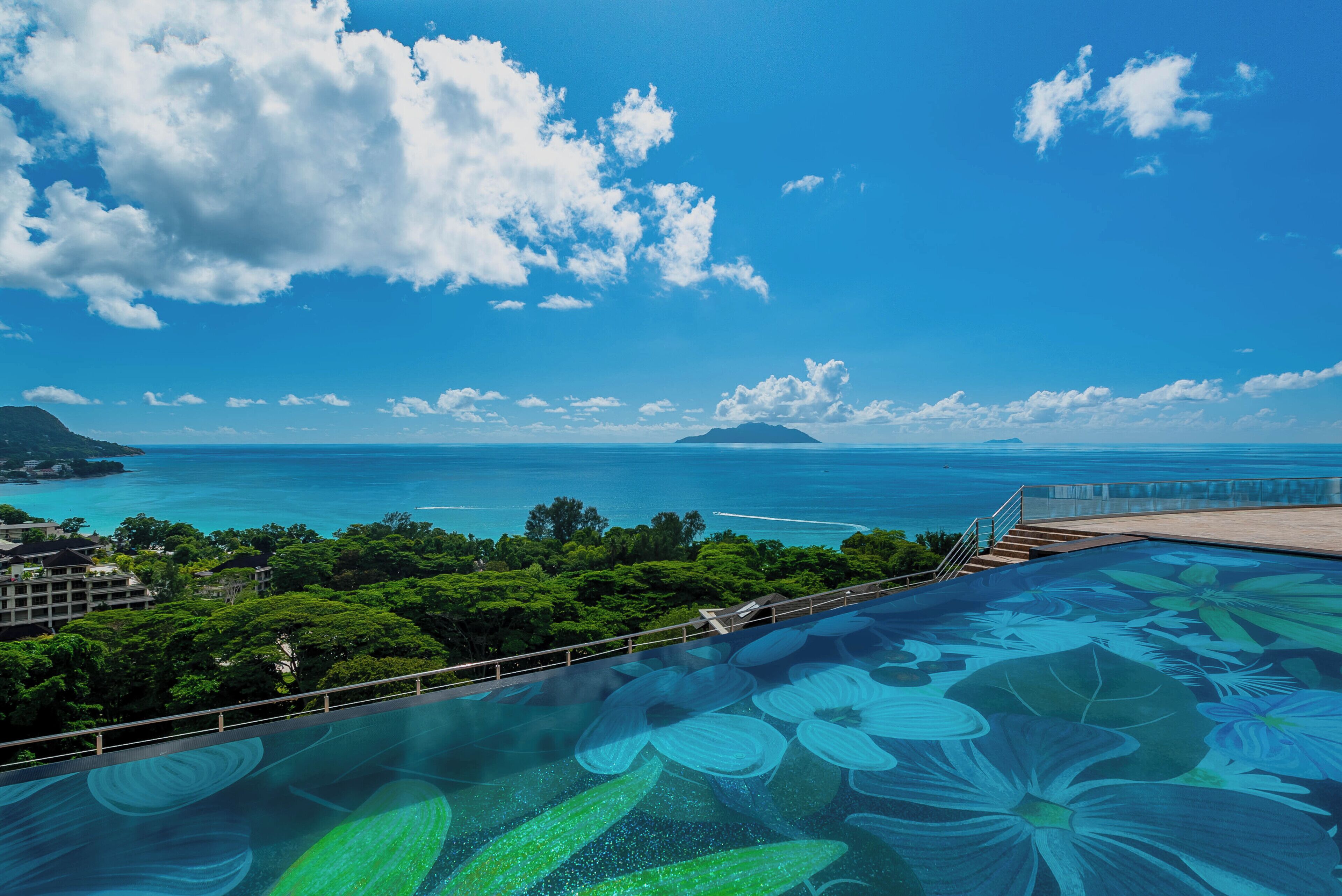 A rooftop infinity pool with a floral design, overlooking a coastal landscape with a clear sky and distant islands.