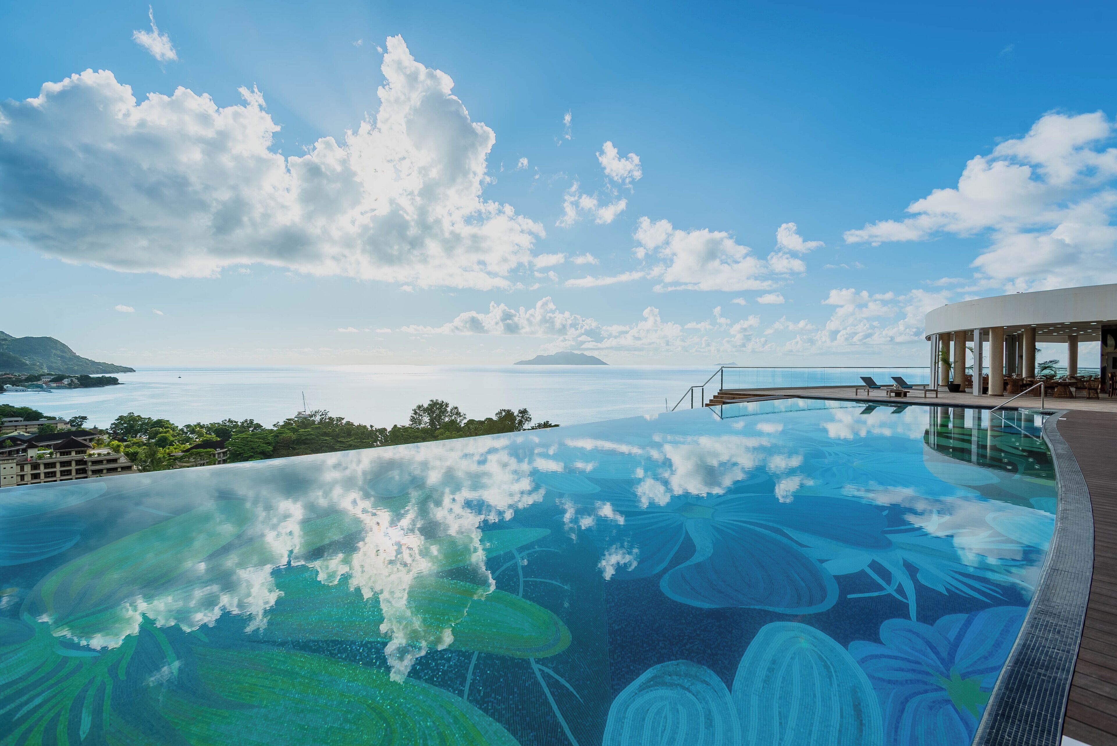 Una piscina infinita con vista al océano y una isla distante, rodeada por un edificio moderno con una terraza cubierta.