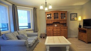 A living room with a striped sofa, a white coffee table, wooden cabinets, and a television. - Apartment in Blankenberge near the Beach (BLANKENBERGE)